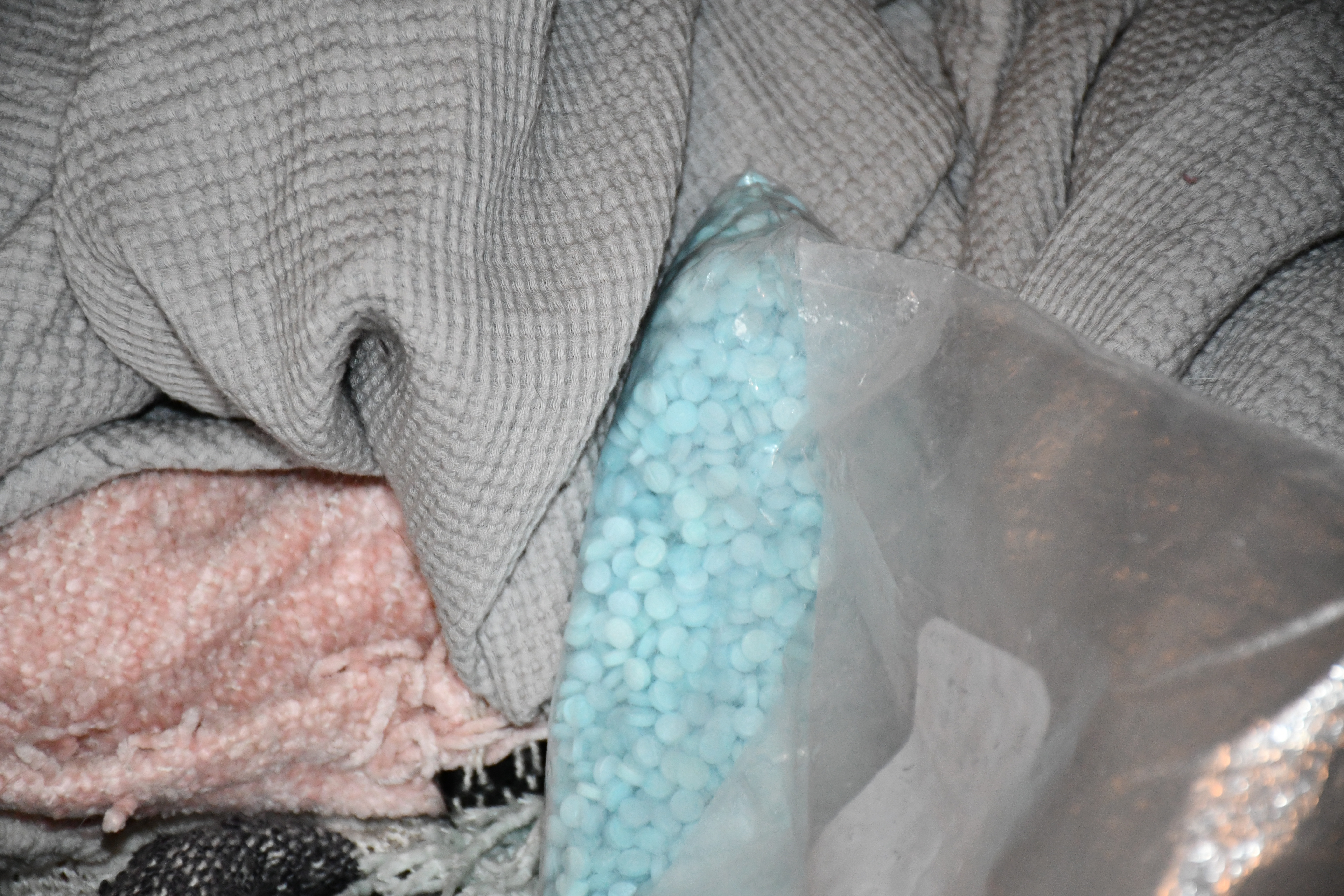 Small blue pills inside a clear plastic bag laying on top of a gray comforter.