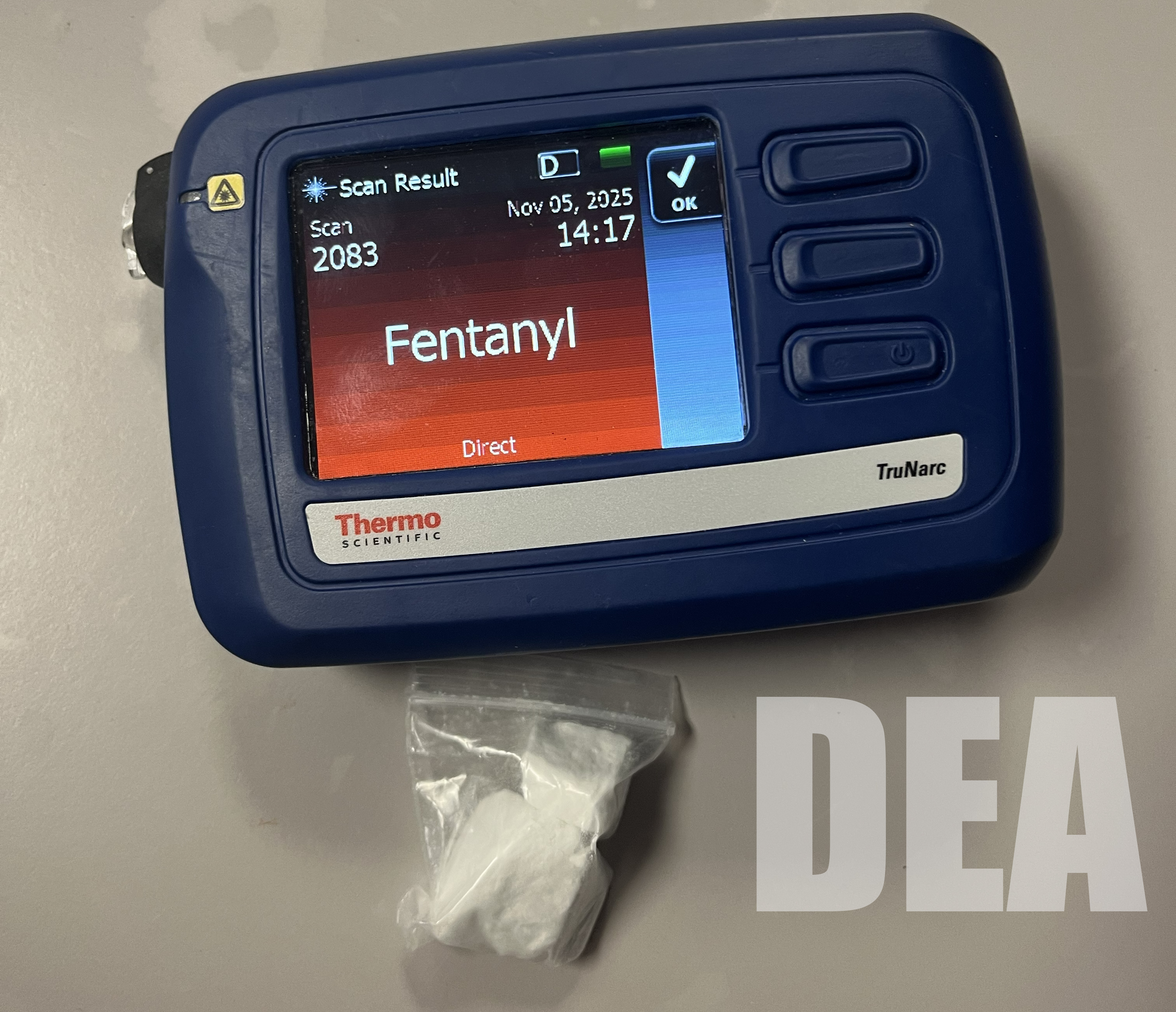 A small brick of white fentanyl powder sits in a clear plastic bag beneath a screen that reads fentanyl.