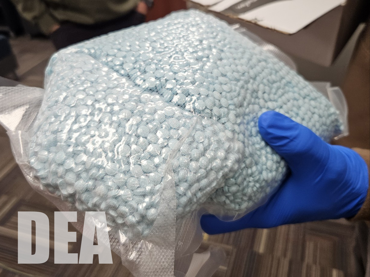 A blue gloved hand holds blue, round pills contained in a clear plastic bag.