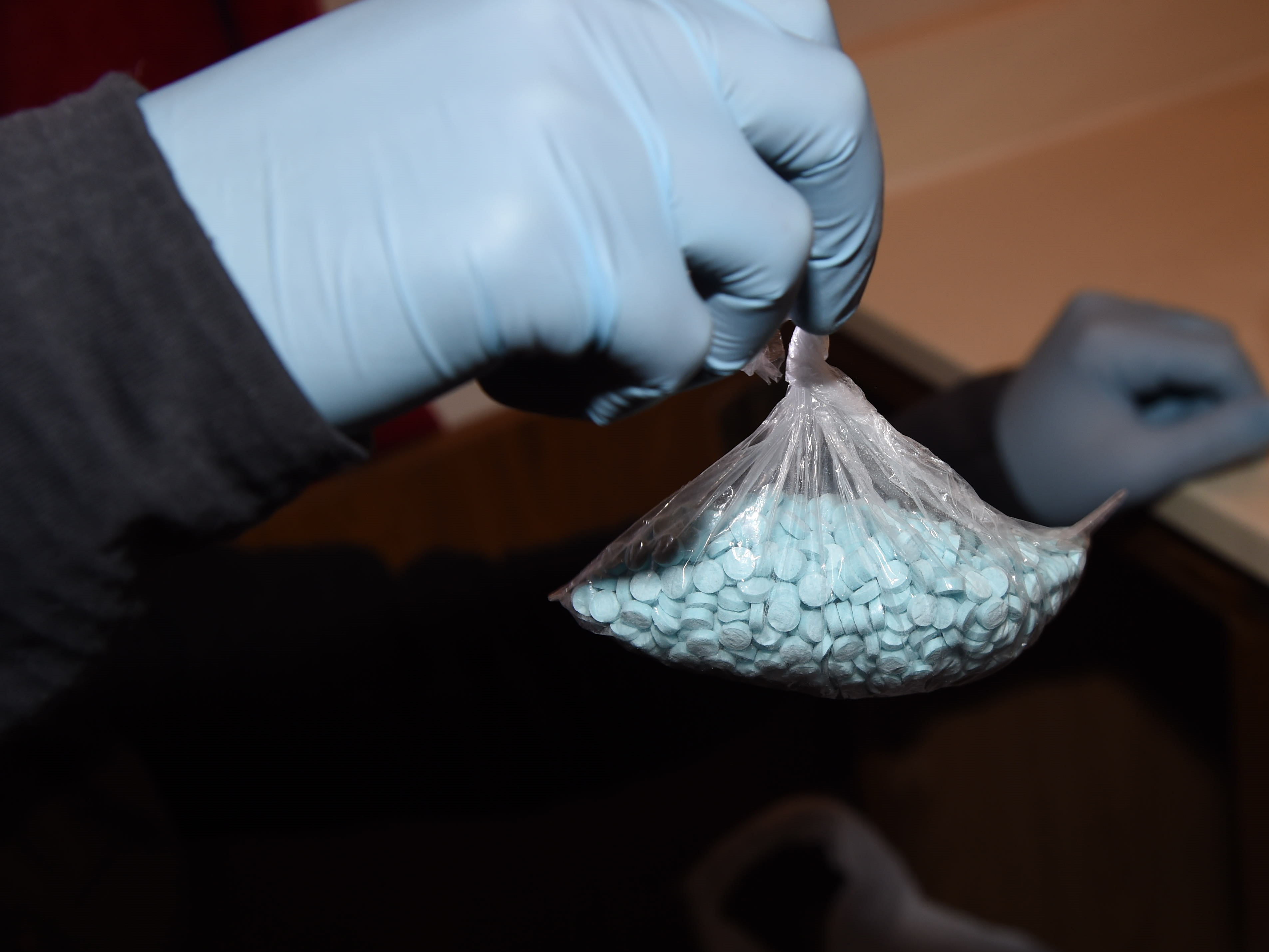 A hand, wearing blue gloves, holds a clear plastic bag containing fentanyl pills.