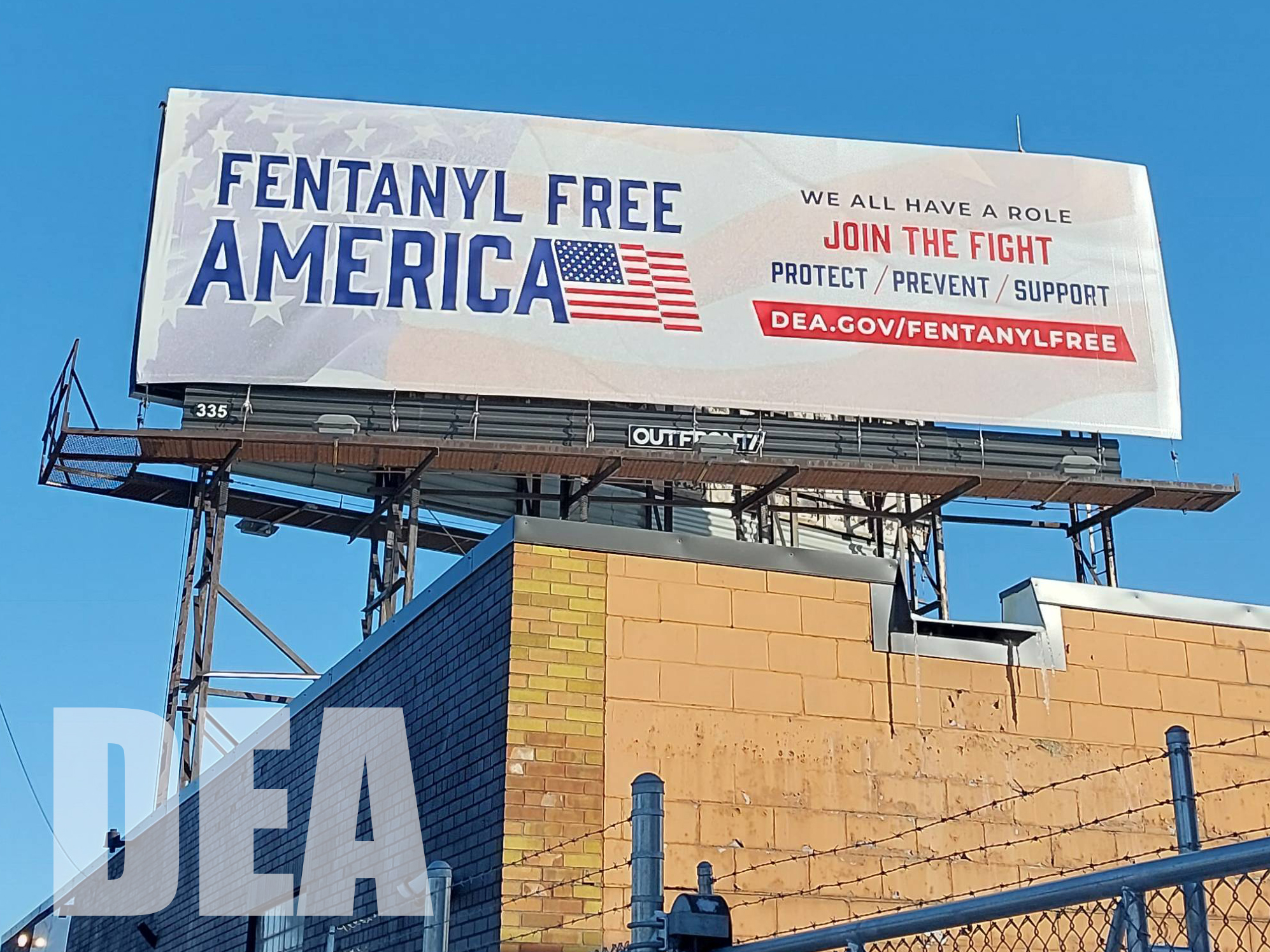 A Fentantyl Free America billbaord with blue text and an American flag stands over a brown building.