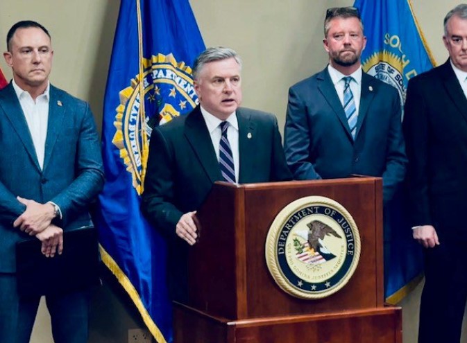 A man in a suit stands behind a podium while three men, also in suits, stand behind him.