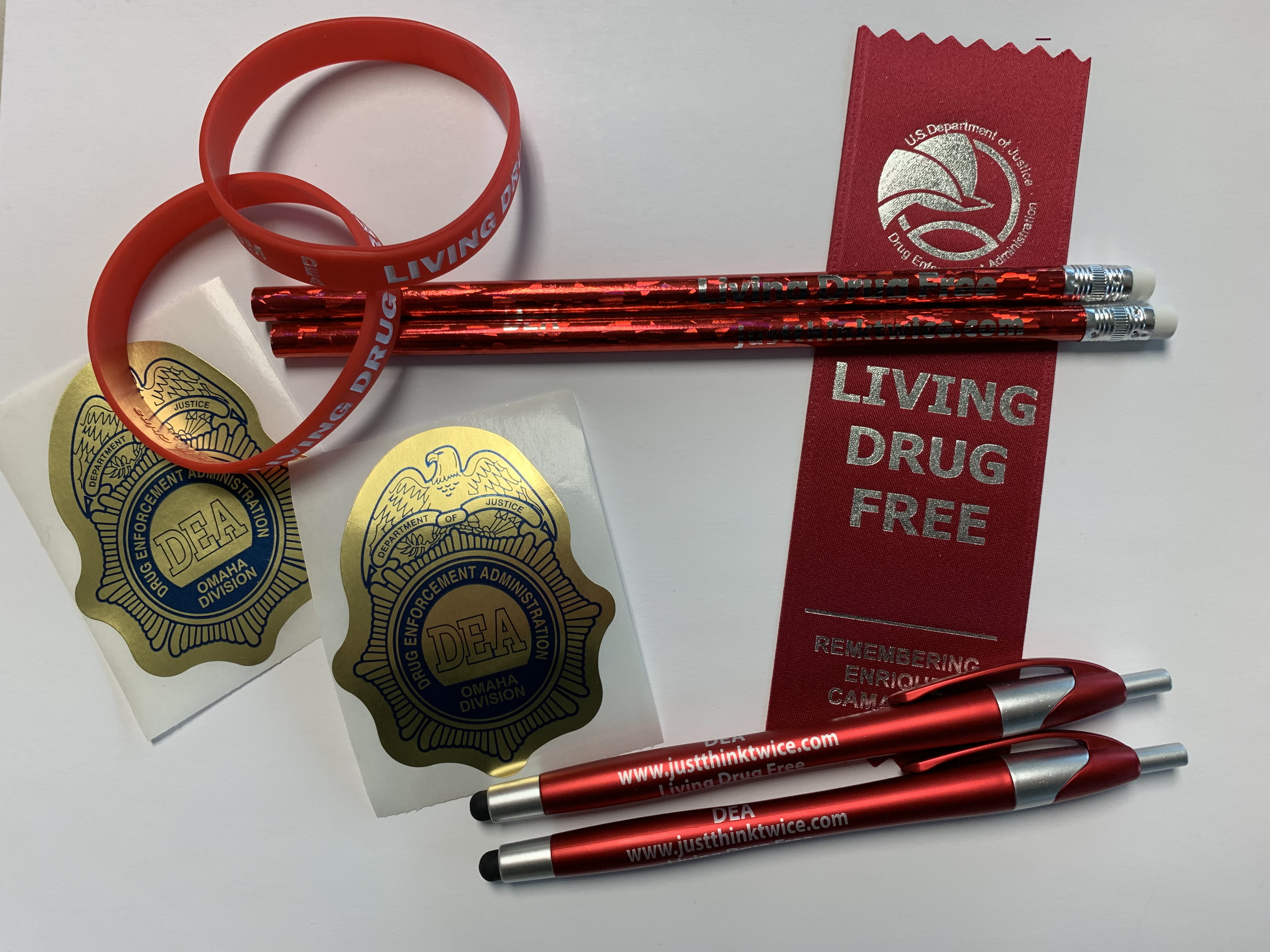 Red bracelets, pencils, pens and a Living Drug Free ribbon sit on a white surface next to DEA badge stickers.