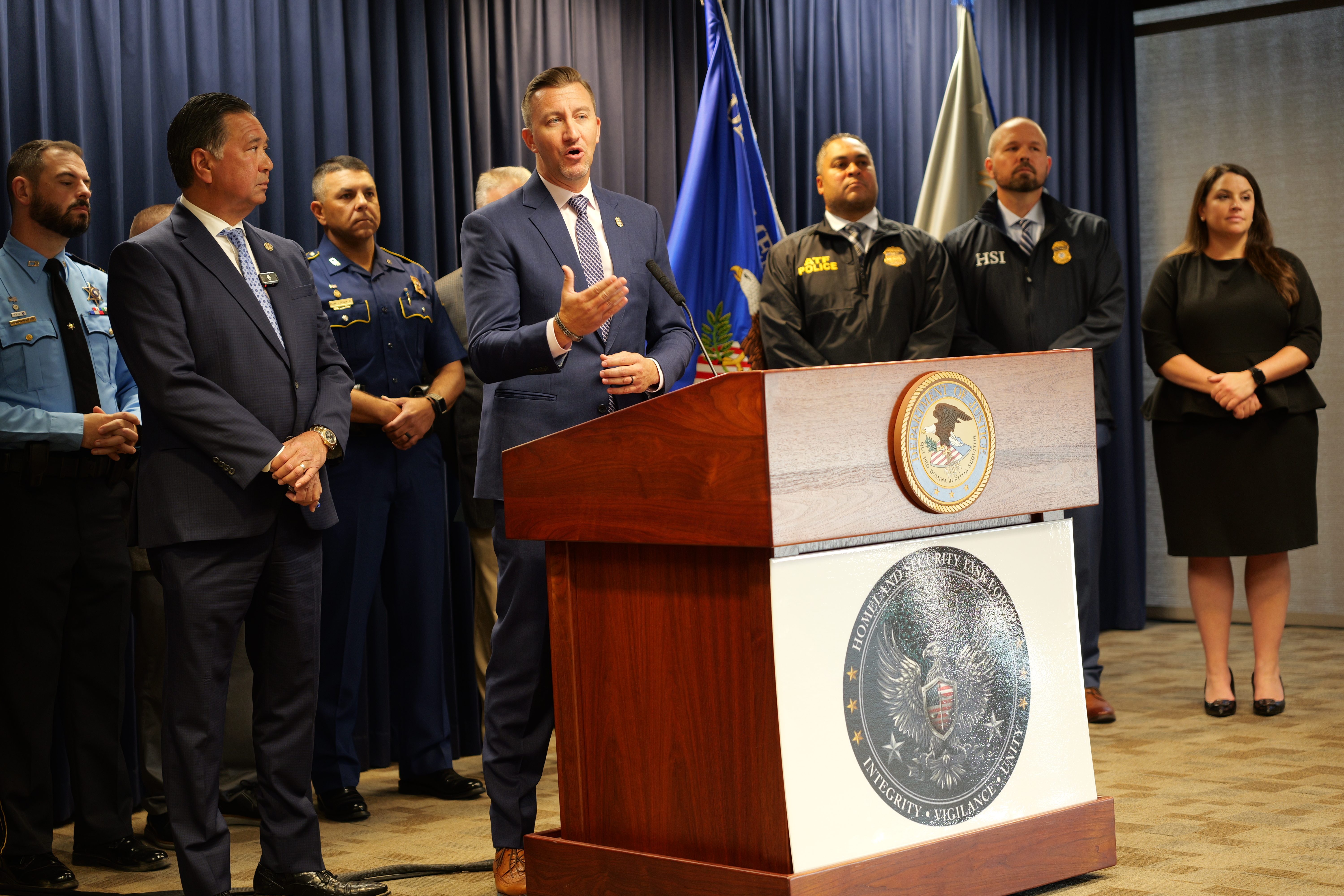 A man wearing a suit speaks at a podium in front of a group of people standing in front of a blue curtain.