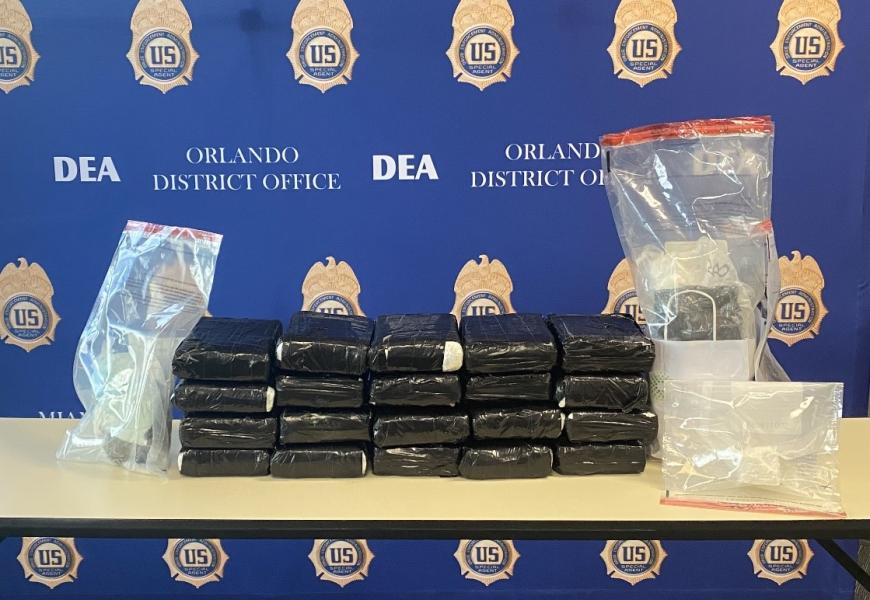 Seized controlled substances contained in clear plastic bags flanking kilograms of controlled substances wrapped in black plastic sitting on a brown table in front of a blue backdrop imprinted with DEA Orlando District Office