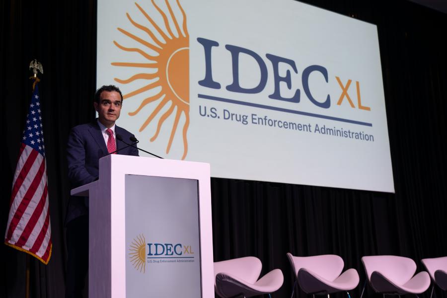 F. Cartwright Weiland, Assistant Secretary of the Bureau of International Narcotics and Law Enforcement Affairs, delivering a presentation at IDEC XL U.S. Drug Enforcement Administration event, with a large logo displayed behi